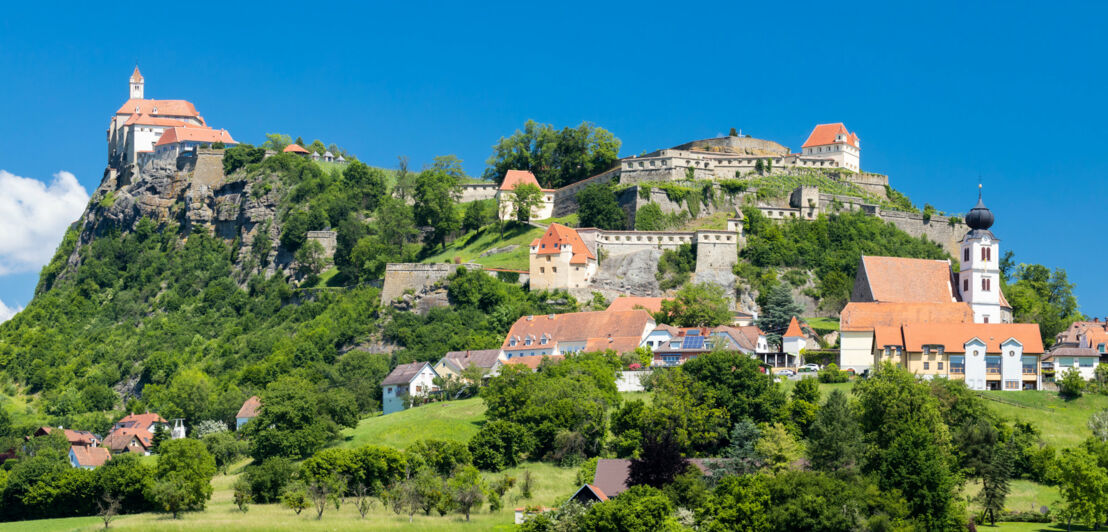 Eine Burganlage mit mehreren Gebäuden auf einem begrünten Felsen unter blauem Himmel.