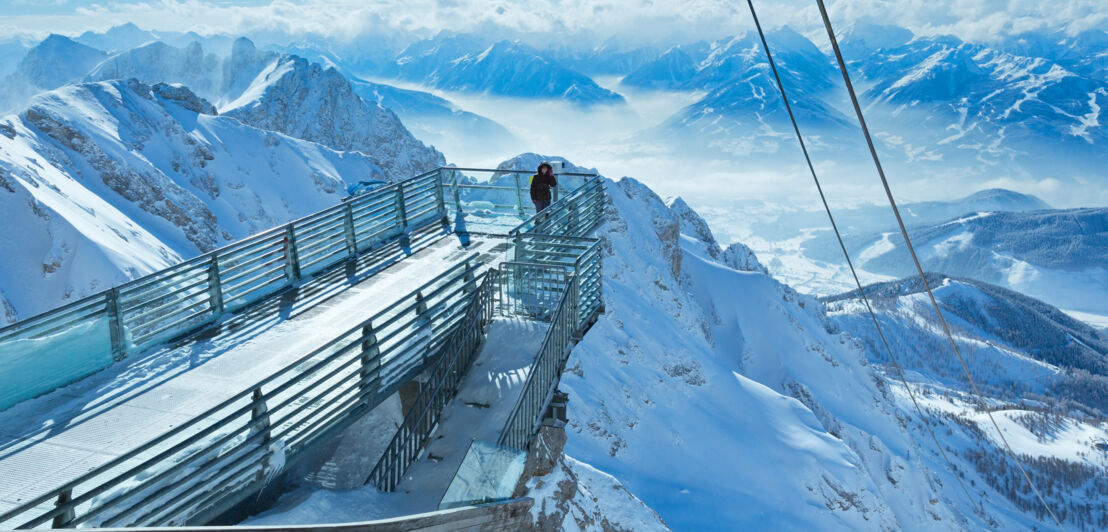 Eine Person steht auf einer Aussichtsplattform vor schneebedecktem Bergpanorama.