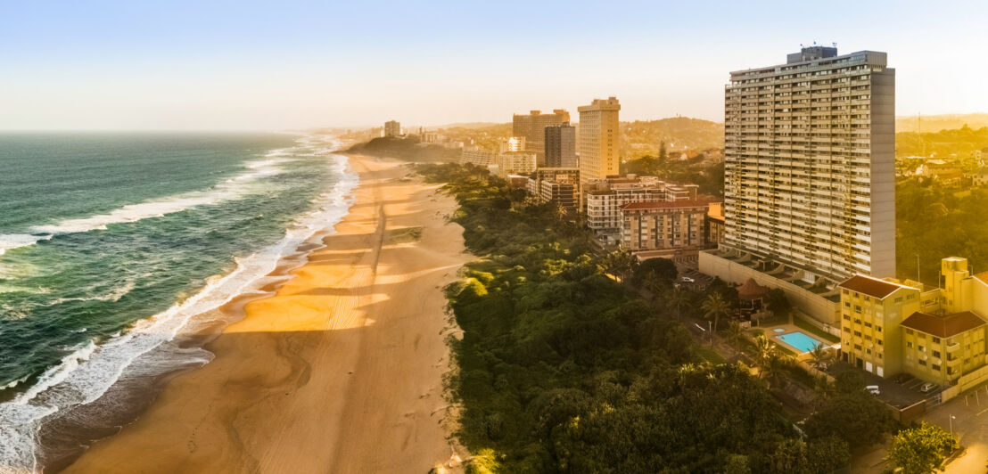 Ein breiter, langer Sandstrand an einer Küste mit großen Hotelbauten im goldenen Sonnenlicht.