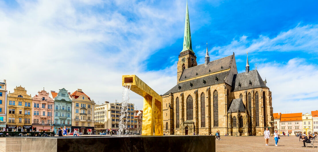 Ein moderner Springbrunnen auf einem Platz mit bunten, historischen Häusern vor einer Kathedrale.