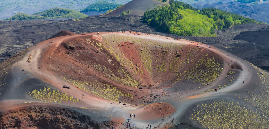 Blick von oben auf einen rotbraunen Krater, drumherum grüne Vegetation. 