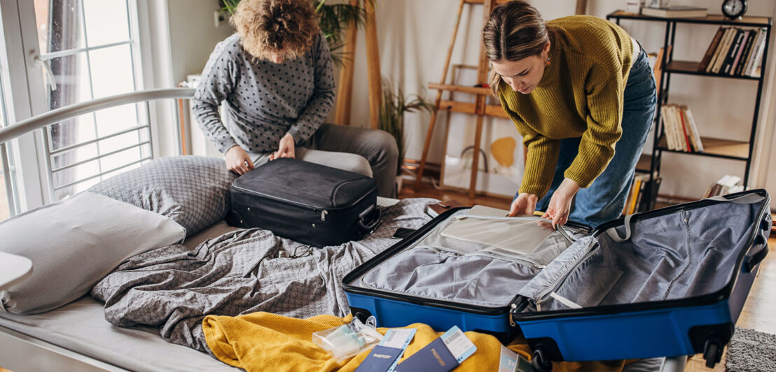 Eine Frau und ein Mann packen gemeinsam jeweils einen Koffer auf einem Bett, Reisepapiere liegen daneben.