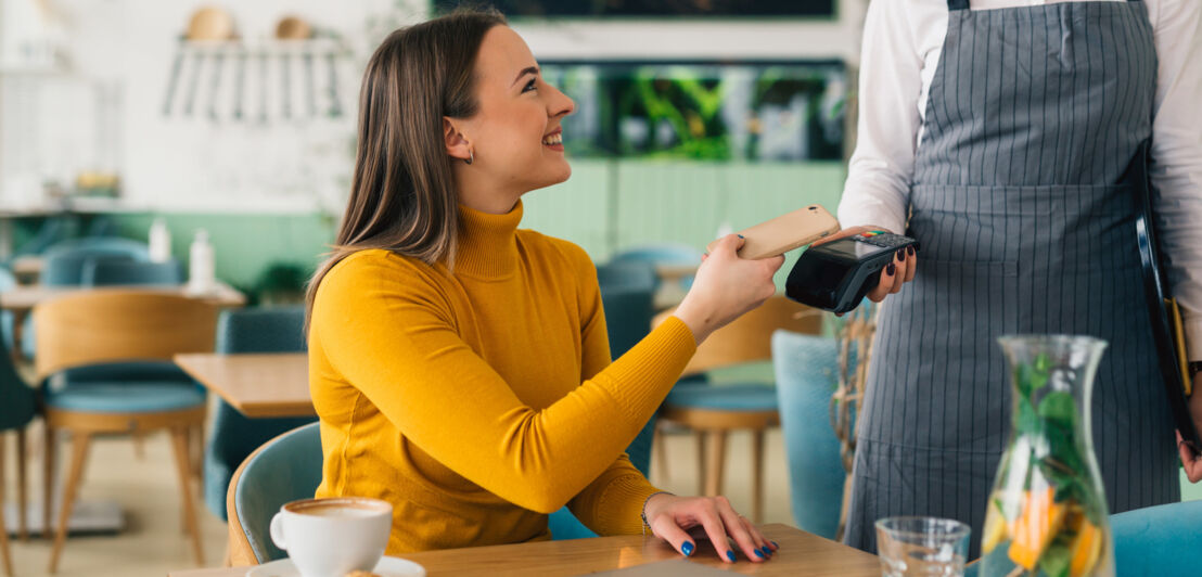 Eine Frau bezahlt kontaktlos mit ihrem Smartphone an einem Tisch in einem Café.