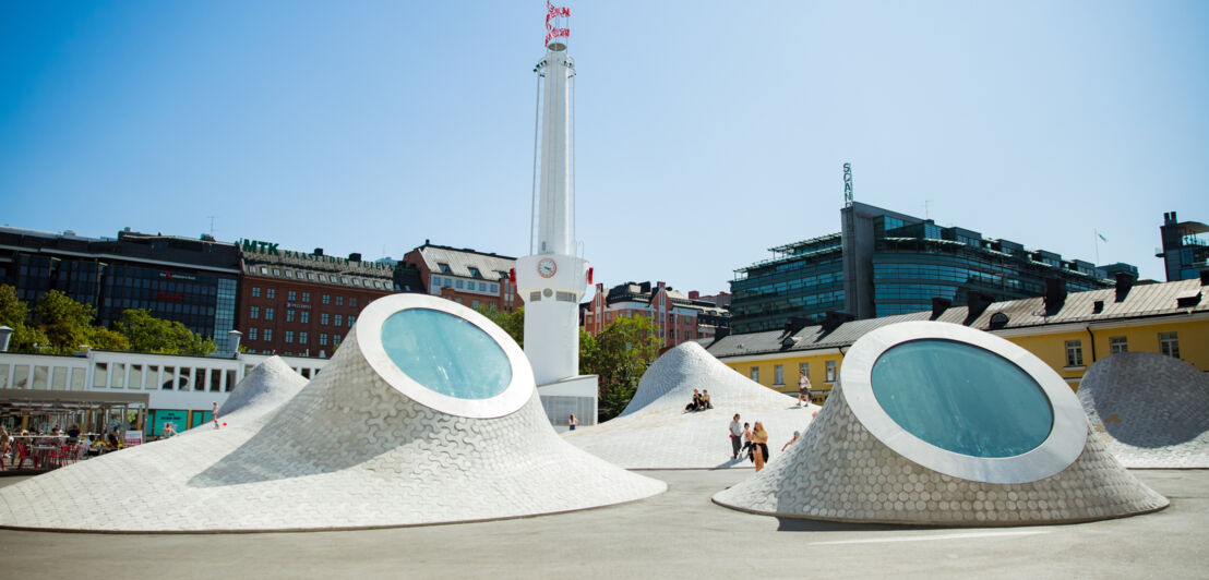 Skulpturenartige Kuppeln mit runden Fenstern auf einem Platz mit Personen in urbaner Umgebung.