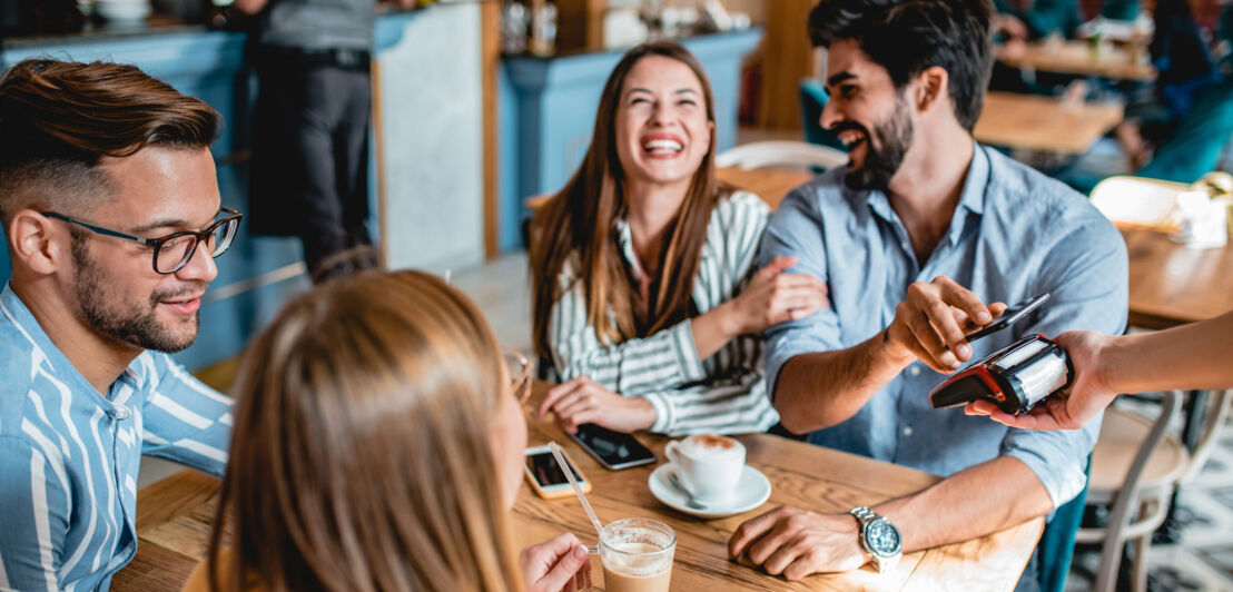 Vier gut gelaunte Personen an einem Tisch in einem Café, eine Person bezahlt kontaktlos mit seinem Smartphone an einem Kartenlesegerät.