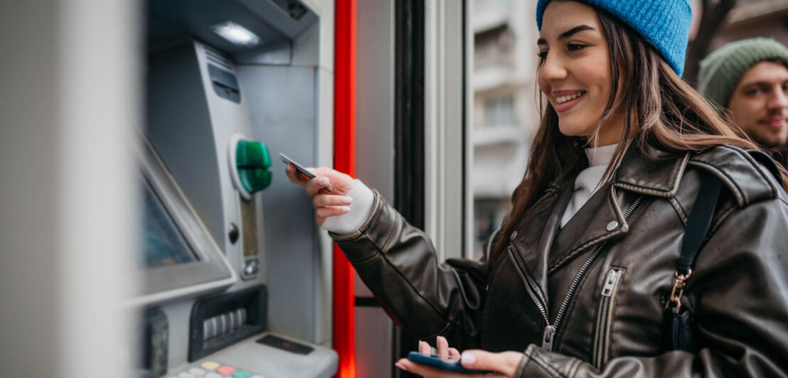 Eine Frau mit Mütze zieht mit einer Kreditkarte Geld an einem Bankautomaten auf der Straße.