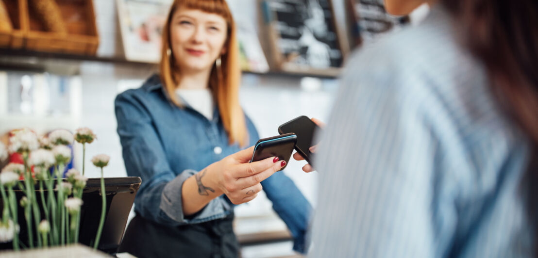 Kontaktlose Zahlung per Smartphone an einem Kartenlesegerät, gehalten von einer Frau hinter einer Theke in einem Geschäft. Kontaktlose Zahlung per Smartphone an einem Kartenlesegerät, gehalten von einer Frau hinter einer Theke in einem Geschäft.