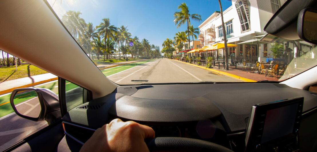 Aufnahme vom Fahrersitz eines Autos mit einer Hand am Lenkrad, das auf einer palmengesäumten Straße fährt Aufnahme vom Fahrersitz eines Autos mit einer Hand am Lenkrad, das auf einer palmengesäumten Straße fährt
