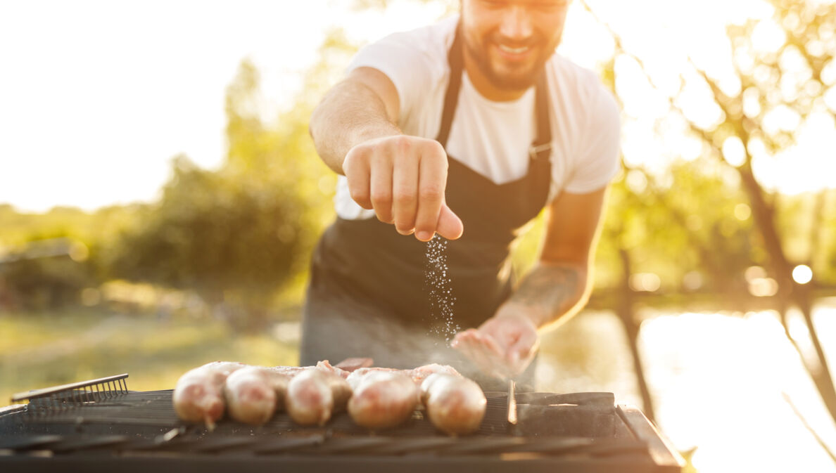 Mann streut Salz auf Würstchen die auf einem Grill liegen