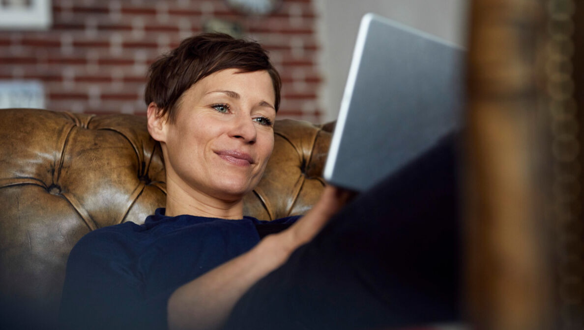 Eine Frau auf einer Ledercouch schaut zufrieden in ihr Tablet