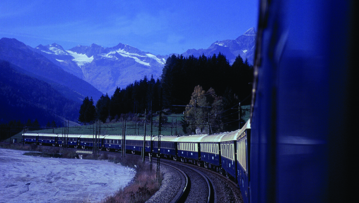 Ein blauer Zug fährt in einer Kurve in der Dämmerung durch die Landschaft, im Hintergrund Wald und Bergpanorama