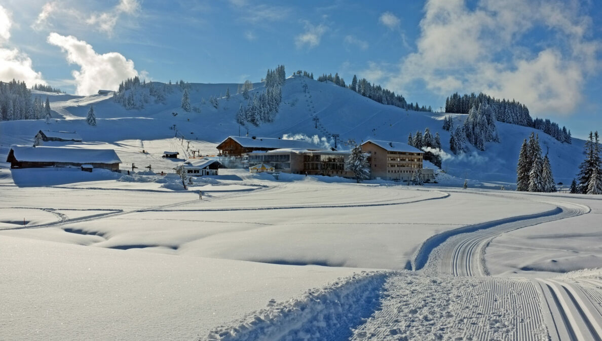 Loipen neben Neuschnee, im Hintergrund Hütten und ein schneebedeckter Berg.