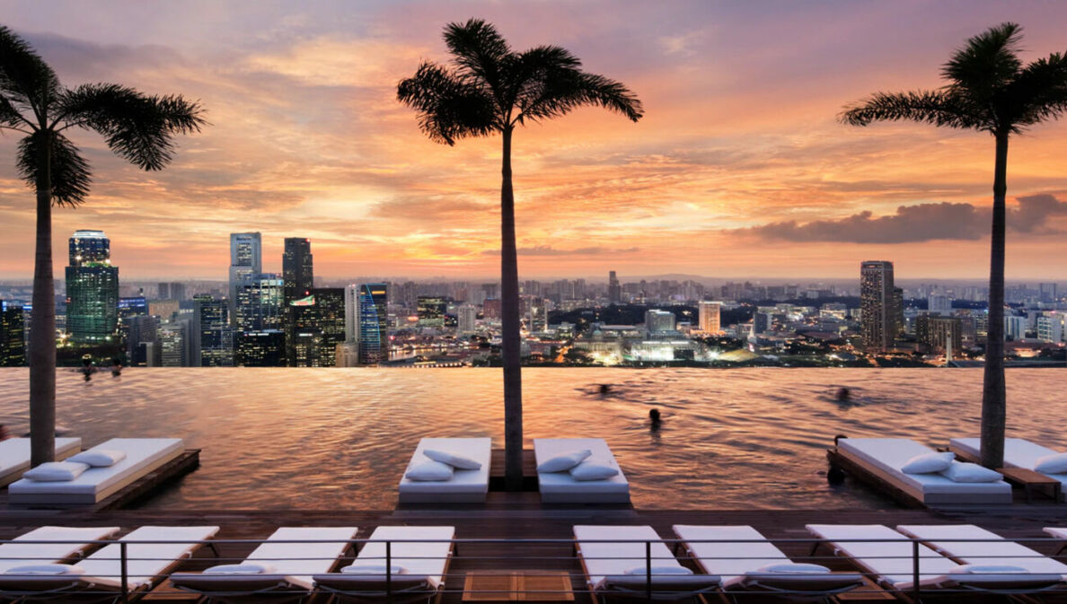 Ein endlos scheinender Pool mit Blick auf die Skyline Singapurs, mit Liegen und Palmen im Wasser