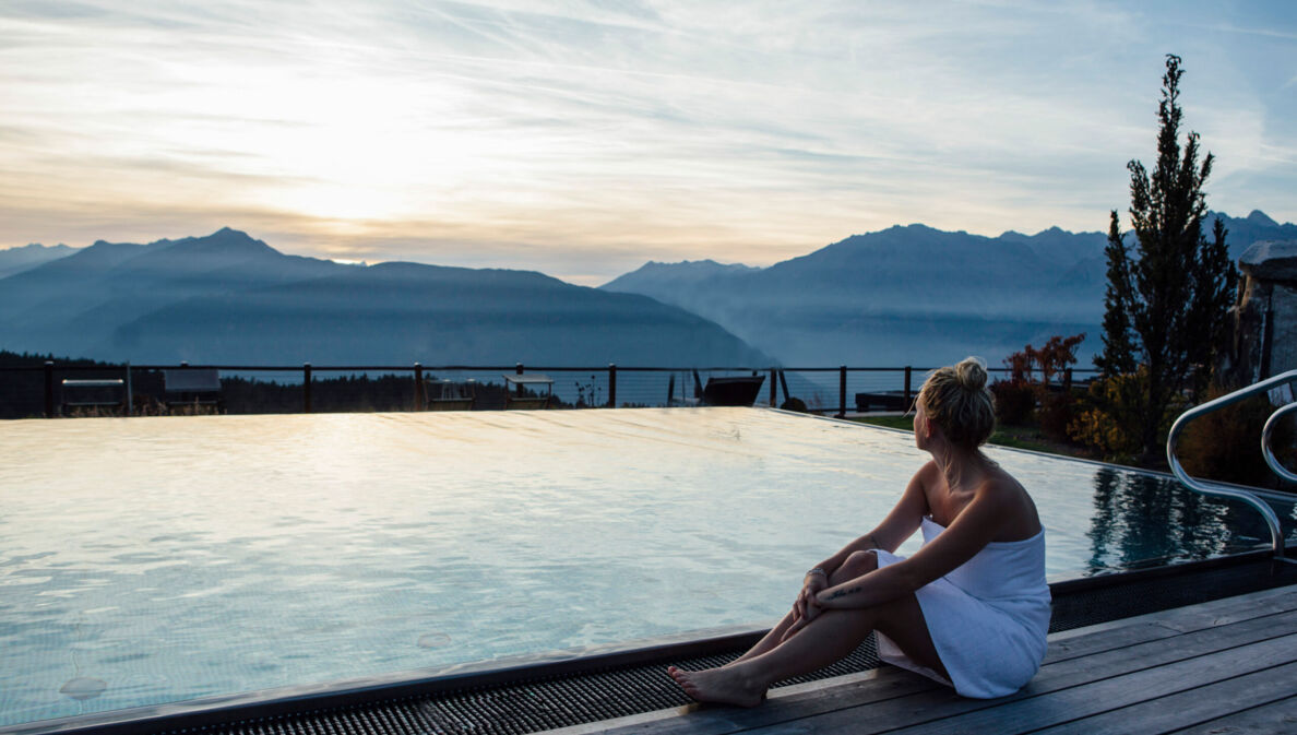 Eine Frau mit umwickelten Handtuch sitzt an einem Outdoor-Pool mit Blick auf die Berge in der Dämmerung