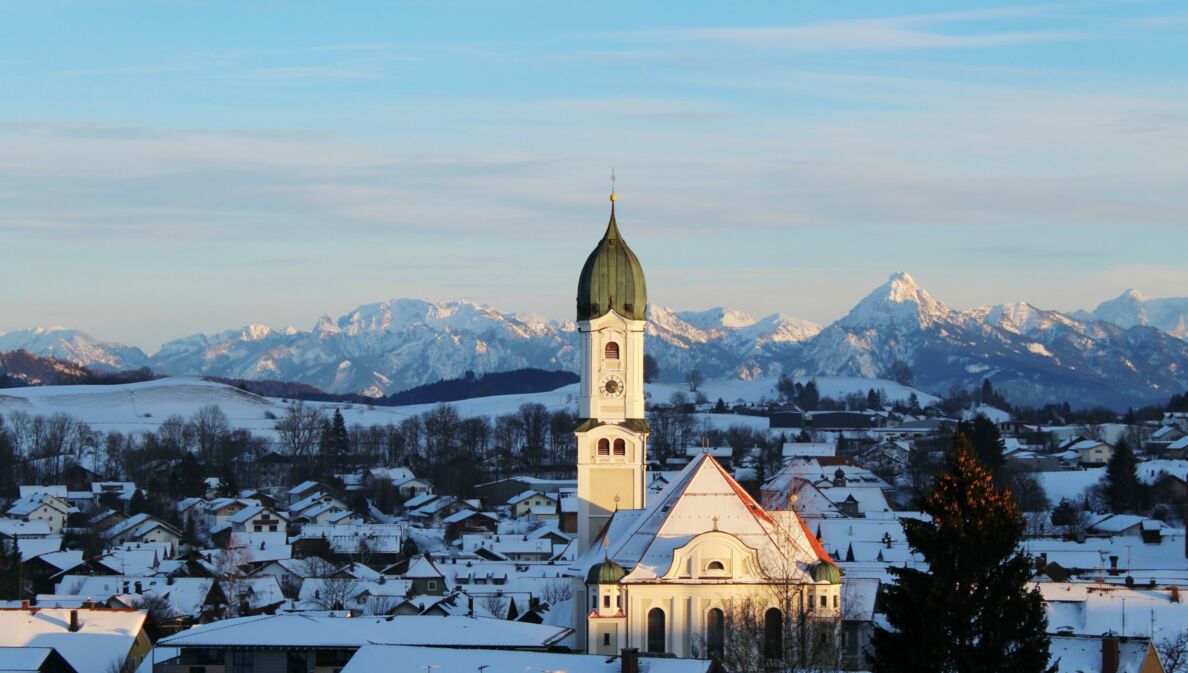 Schneebedeckter Ort mit weißer Kirche vor Alpenpanorama in der Dämmerung