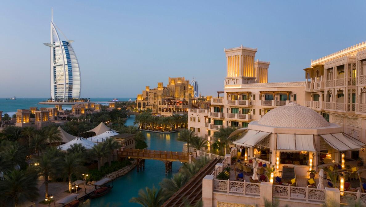 Stadtteil von Dubai mit Gebäuden an einem Fluss in der Abenddämmerung, im Hintergrund das Hotel Burj Al Arab in Segelform am Wasser