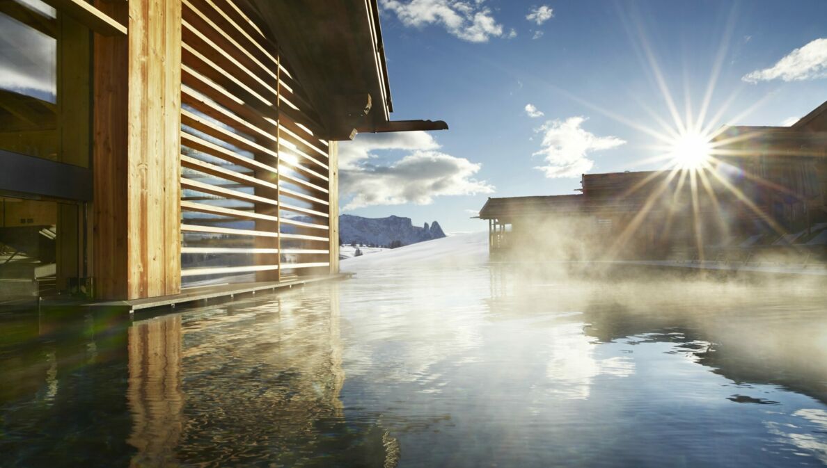 Ein Pool in der strahlenden Sonne, daneben Holzhäuser und Berge im Hintergrund