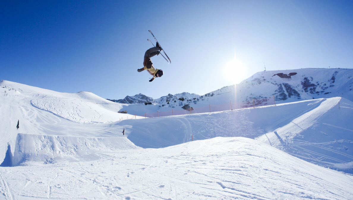 Eine Person, die mit Ski einen Salto in der Luft macht, im Hintergrund ein schneebedeckter Berg