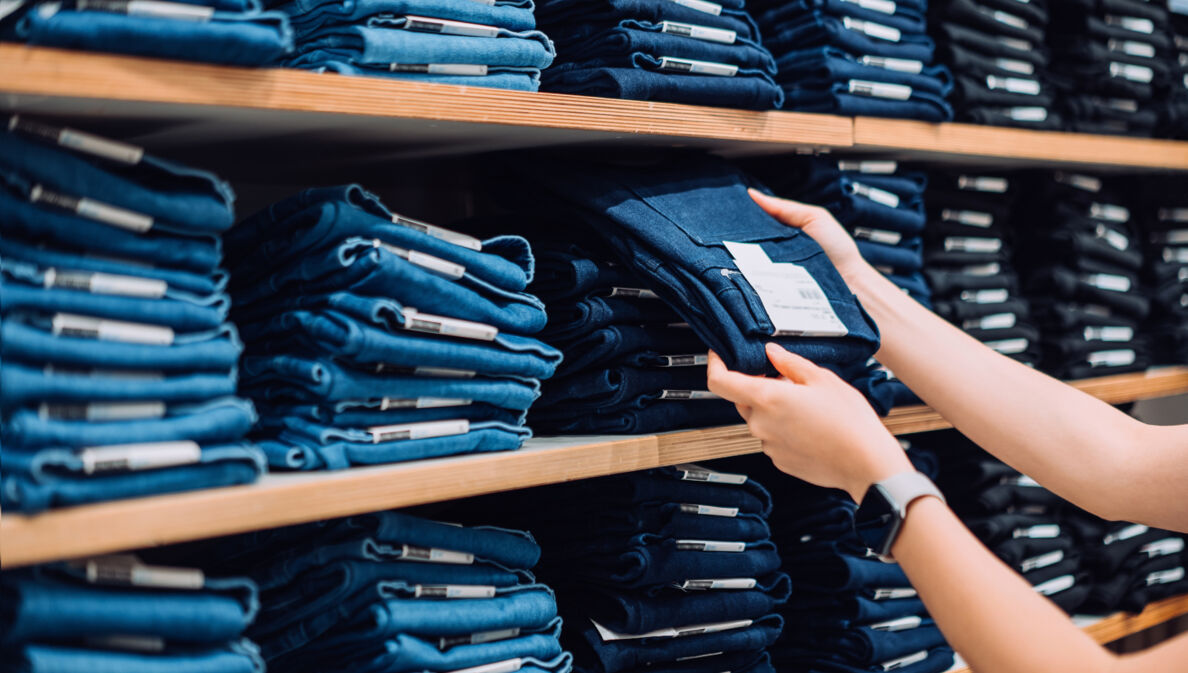 Zwei Hände greifen eine blaue Jeanshose aus einem Regal mit gestapelten Jeanshosen in einem Geschäft