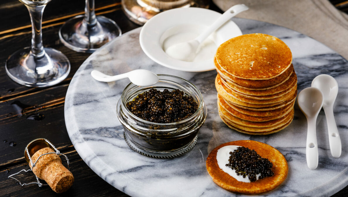 Kaviar im Glas, ein Stapel Blinis und Löffel auf einem Marmortisch, daneben Sektkorken und -gläser