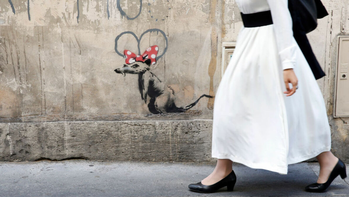 Eine Frau in langem, weißen Kleid läuft an einem Banksy-Werk vorbei: eine Ratte mit roter Minnie-Mouse-Schleife auf dem Kopf.