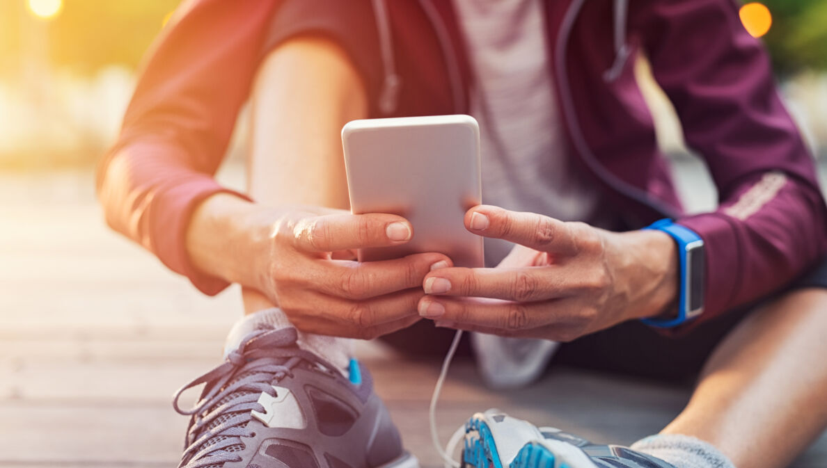 Sportlich gekleidete Person sitzt auf dem Boden und hält ein Handy in der Hand.