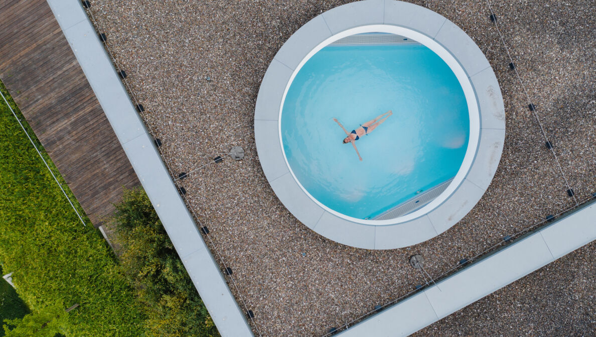 Eine Frau schwebt mit ausgestreckten Armen in einem Pool, fotografiert durch ein rundes Dachfenster aus der Vogelperspektive