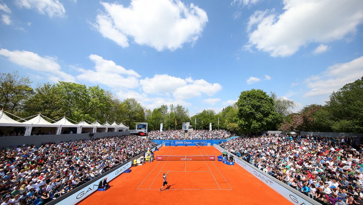 Tennisspieler spielen auf einem Sandplatz, umgeben von zahlreichen Zuschauern.