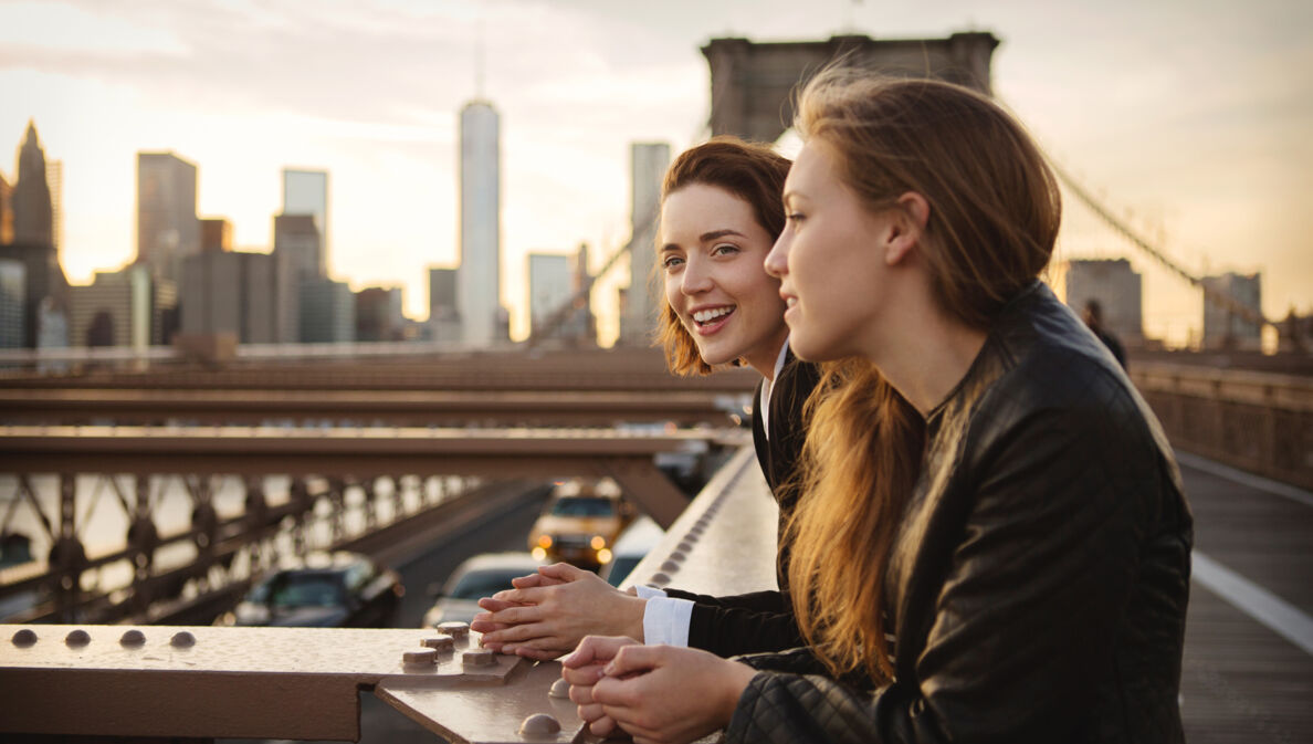 Zwei junge Mädchen stehen lächelnd auf der Brooklyn Bridge vor der Skyline Manhattans