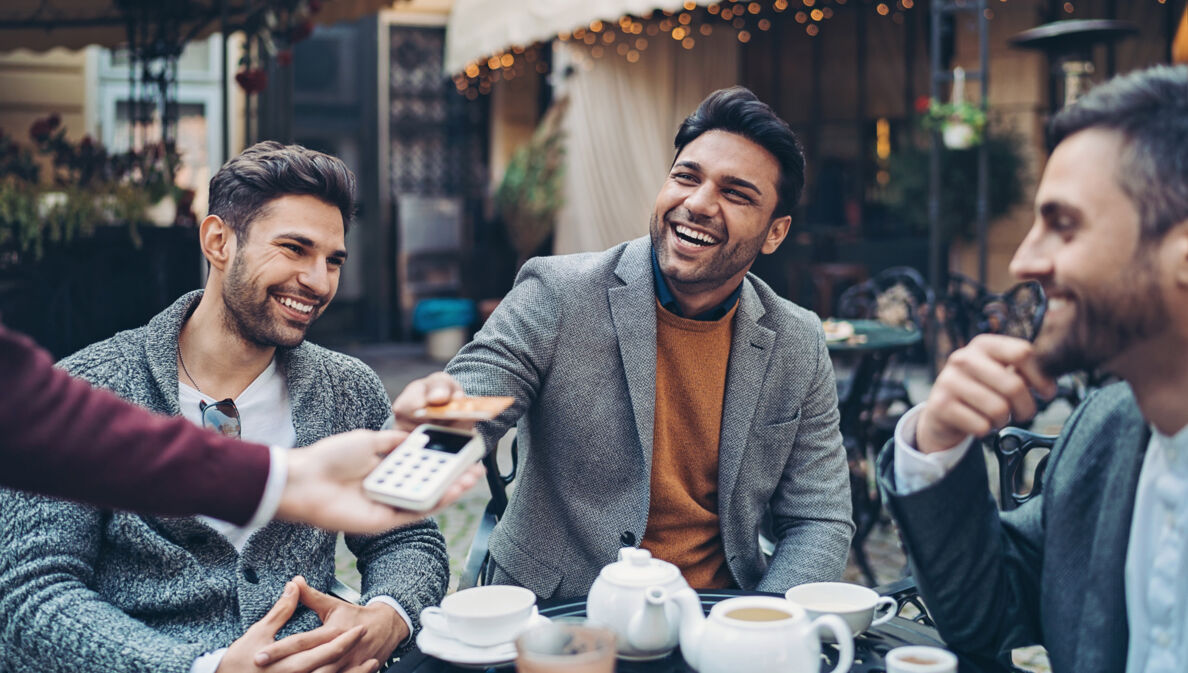 Ein junger, gut gelaunter Mann bezahlt kontaktlos mit seiner American Express Gold Card im Beisein seiner Freunde an einem Tisch in einem Café 