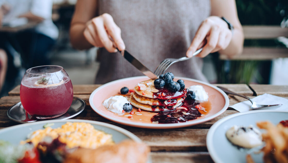 Eine Person sitzt an einem gedeckten Tisch vor einem Teller mit Pancakes im Außenbereich eines Cafés