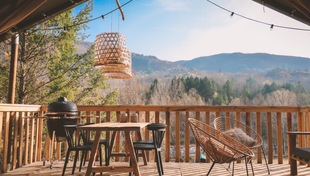 Holzterrasse mit Tisch, Stühlen und Lampen, im Hintergrund bewaldete Hügel