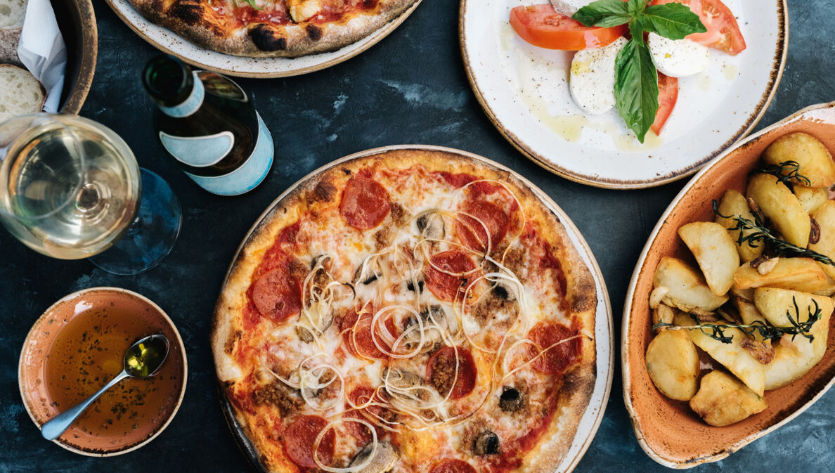 Aufsicht einer Pizza neben weiteren italienischen Gerichten und einem Glas Wein auf einem Tisch