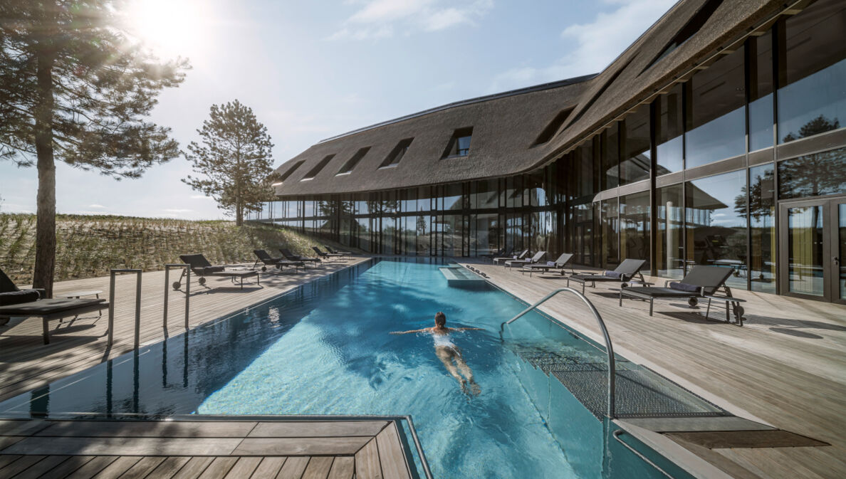 Ein luxuriöses Hotel in den Dünen mit Reetdach und Außenpool, in dem eine Frau schwimmt