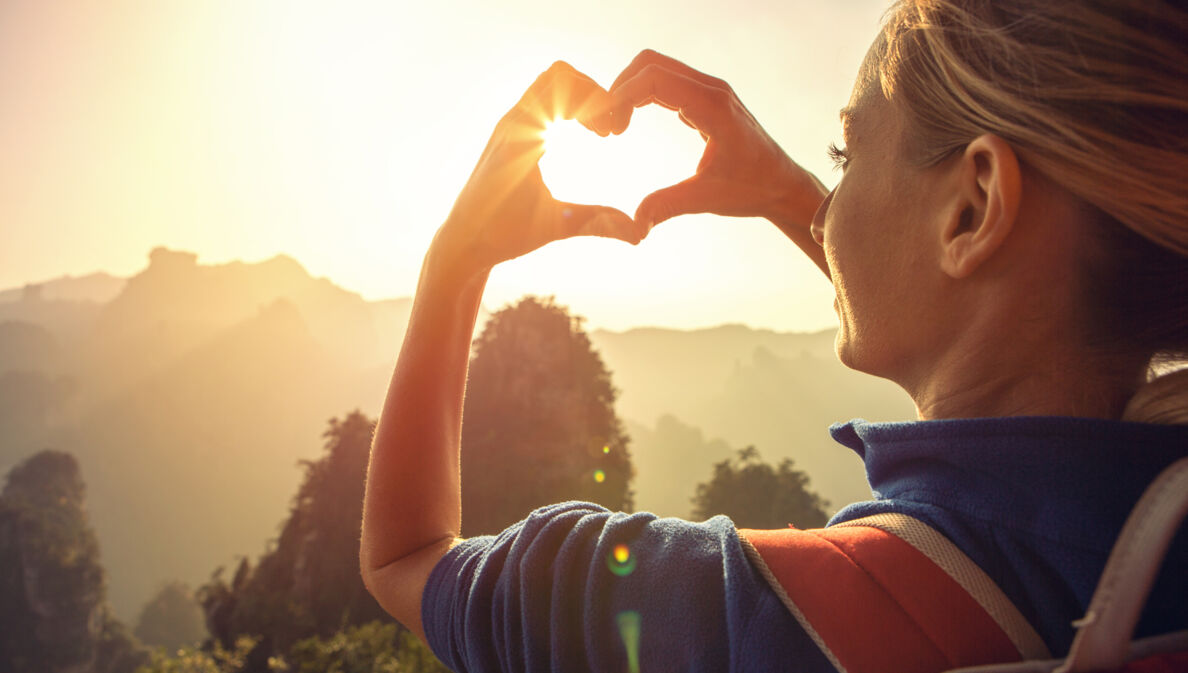 Eine junge Frau blickt in die Natur und formt ihre Hände zu einem Herz, durch das die Sonne scheint