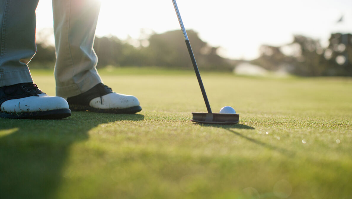 Nahaufnahme von einem Golfschläger, dahinter ein weißer Golfball auf einem grünen Rasen