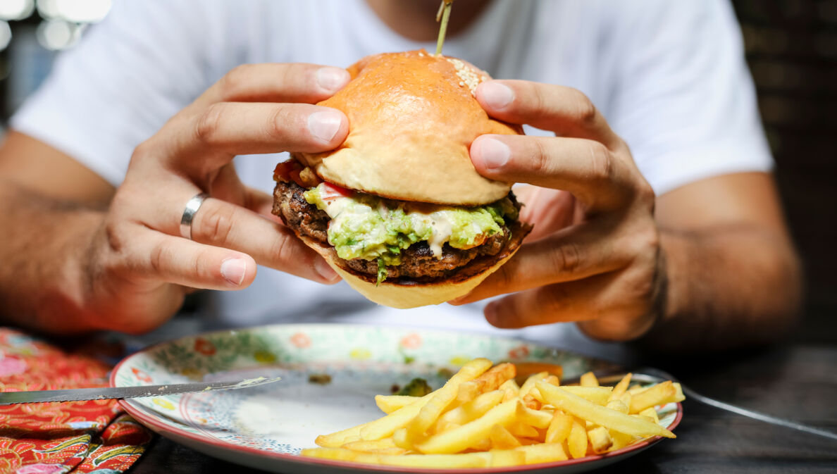 Eine Person hält einen Burger mit beiden Händen über einem Teller mit Pommes fest