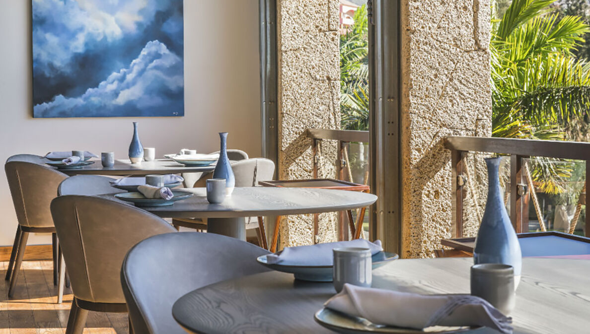 Innenraum eines luftigen, hellen Restaurants mit gedeckten Tischen an einer Fensterfront mit Blick auf Palmen