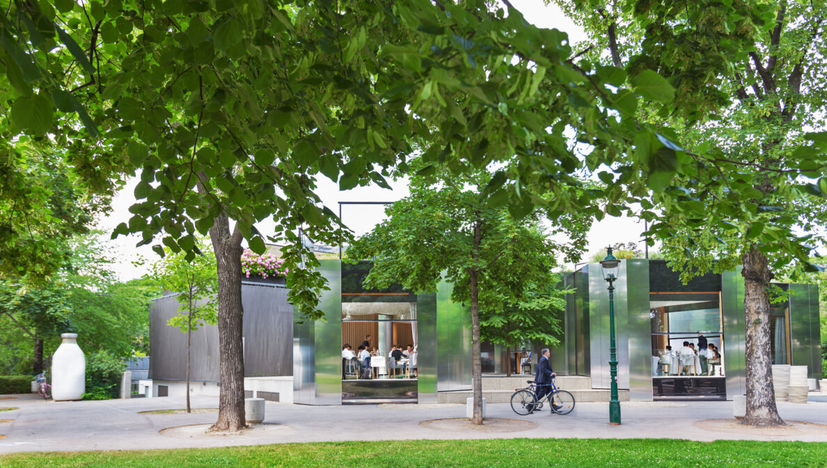 Rendering des Wiener Stadtparks mit Bäumen und Wegen, im Hintergrund ein Restaurant mit offenen Türen und Gästen im Inneren