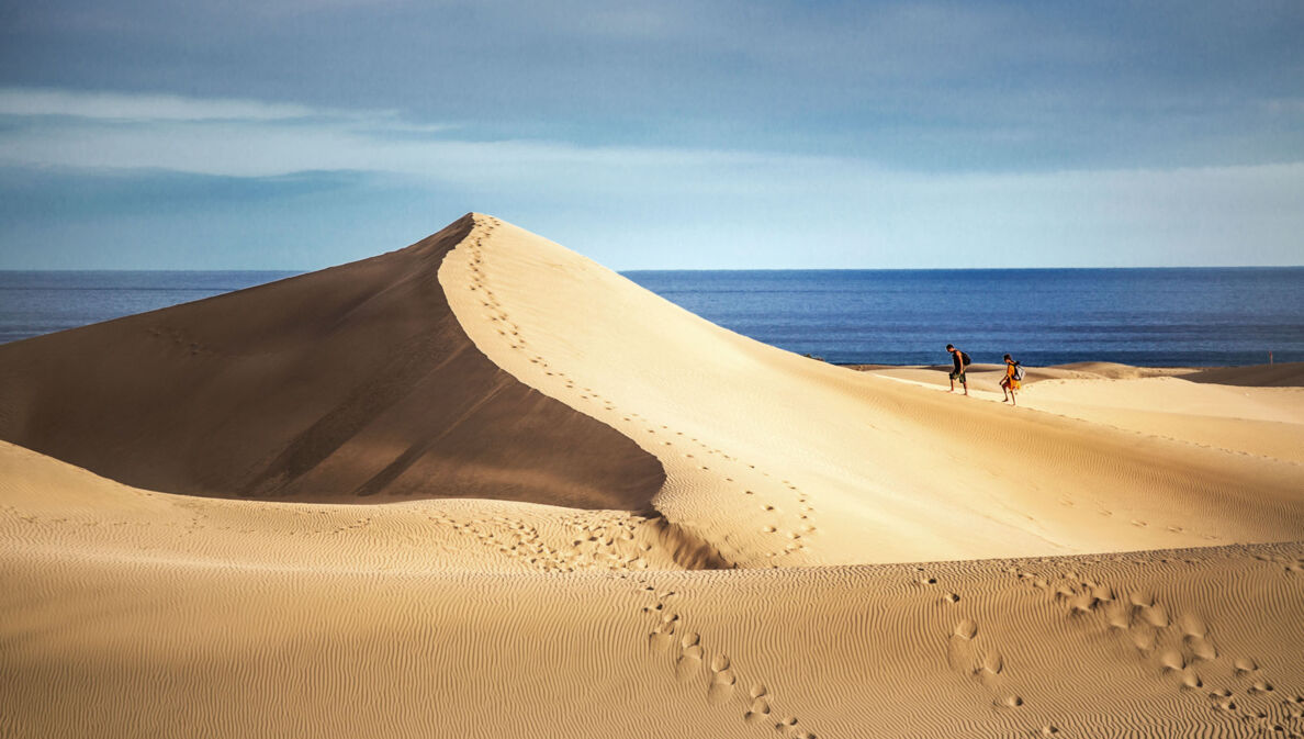 Zwei Personen wandern auf einer großen Sanddüne am Meer