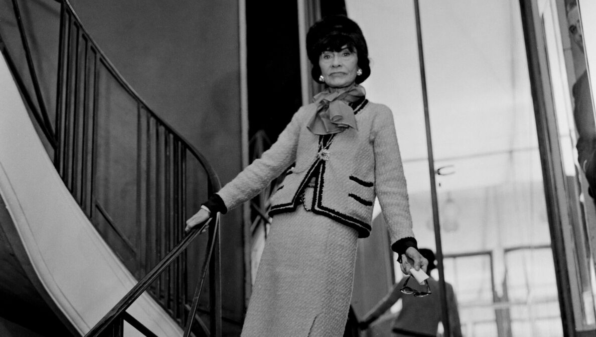 Schwarzweißaufnahme von Coco Chanel in einem Tweed-Kostüm auf einer Treppe vor einer Spiegelwand