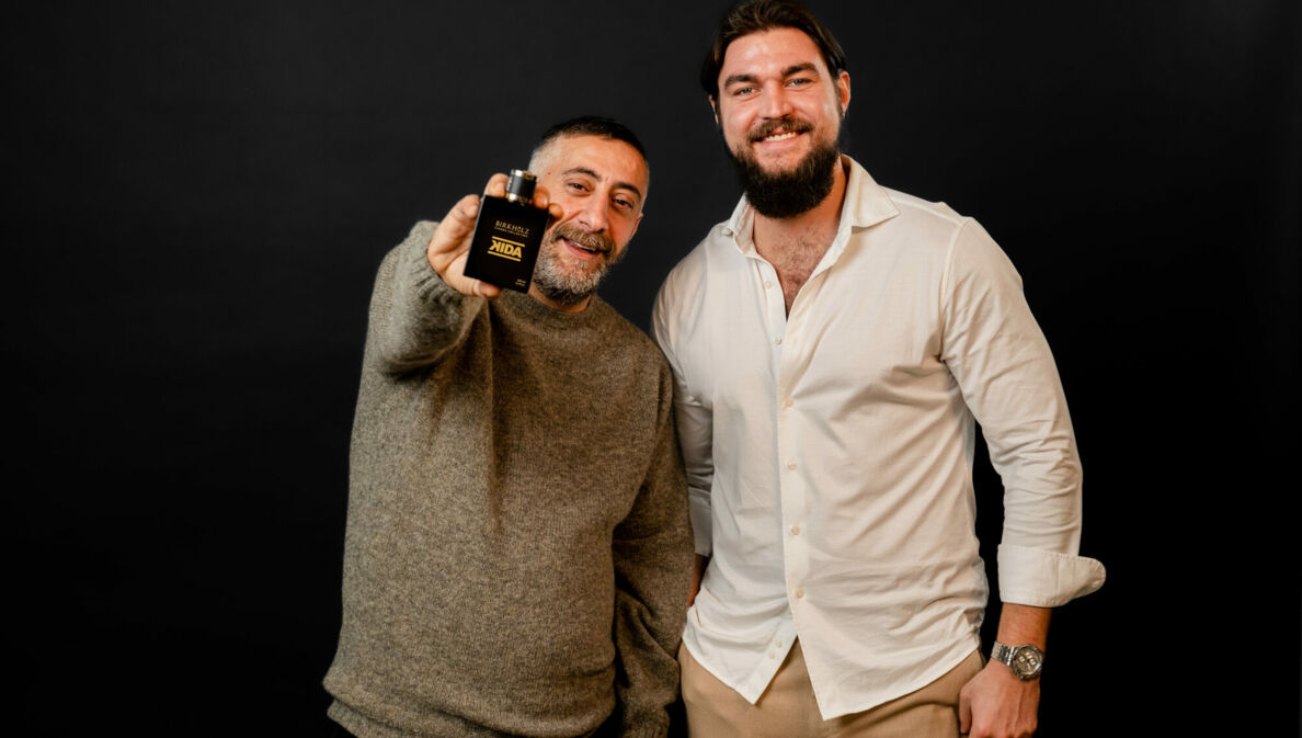 Kida Khodr Ramadan und Philip Birkholz stehen Schulter an Schulter vor einem schwarzen Hintergrund. Kida Khodr Ramadan hält den Flakon des Parfüms KIDA in die Kamera