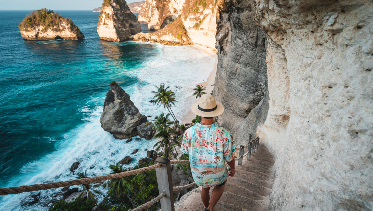 Rückansicht eines Mannes mit Strohhut und Hawaiihemd, der eine schmale Steintreppe an einer Felswand hinunter zu einer palmengesäumten Strandbucht geht