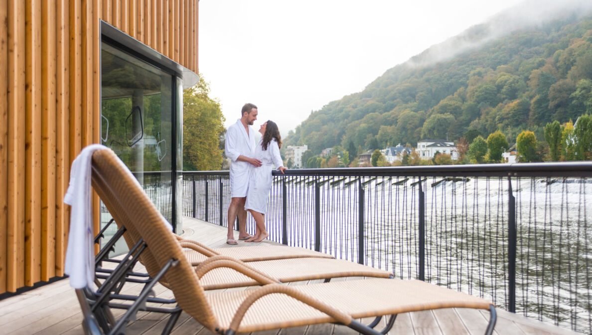 Ein Mann und eine Frau in weißen Bademänteln stehen auf dem Sonnendeck einer Flusssauna