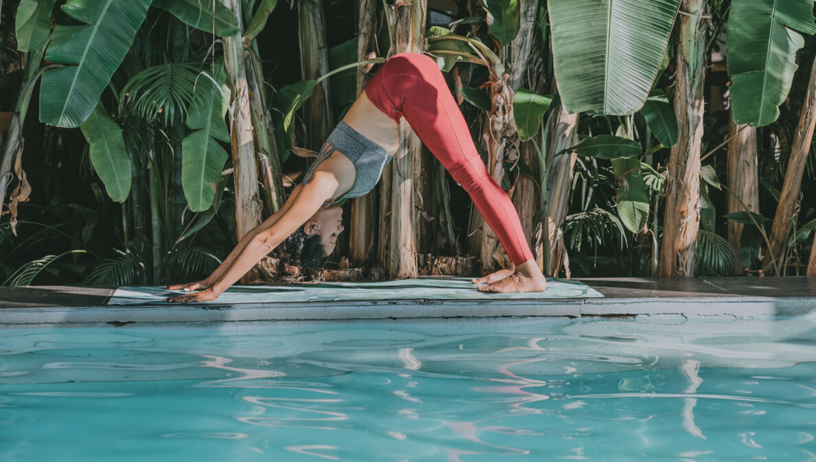 Eine junge Frau bei einer Yogaübung auf einer Matte an einem Pool, umgeben von Palmen