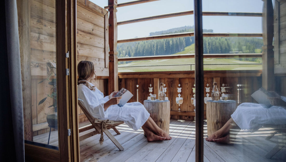 Eine Frau in weißem Bademantel sitzt mit einem Buch auf einer Terrasse eines Holzchalets mit Blick in die Berge