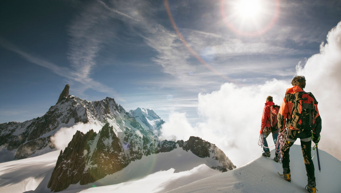 Zwei Personen gehen einen schneebedeckten Bergkamm entlang, im Hintergrund erhebt sich ein noch höheres Bergpanorama