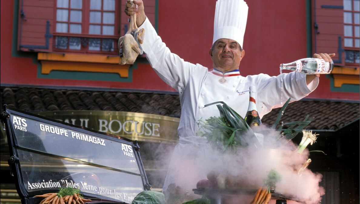 Paul Bocuse in Kochuniform bei einer Inszenierung mit Lebensmitteln vor seinem Restaurant