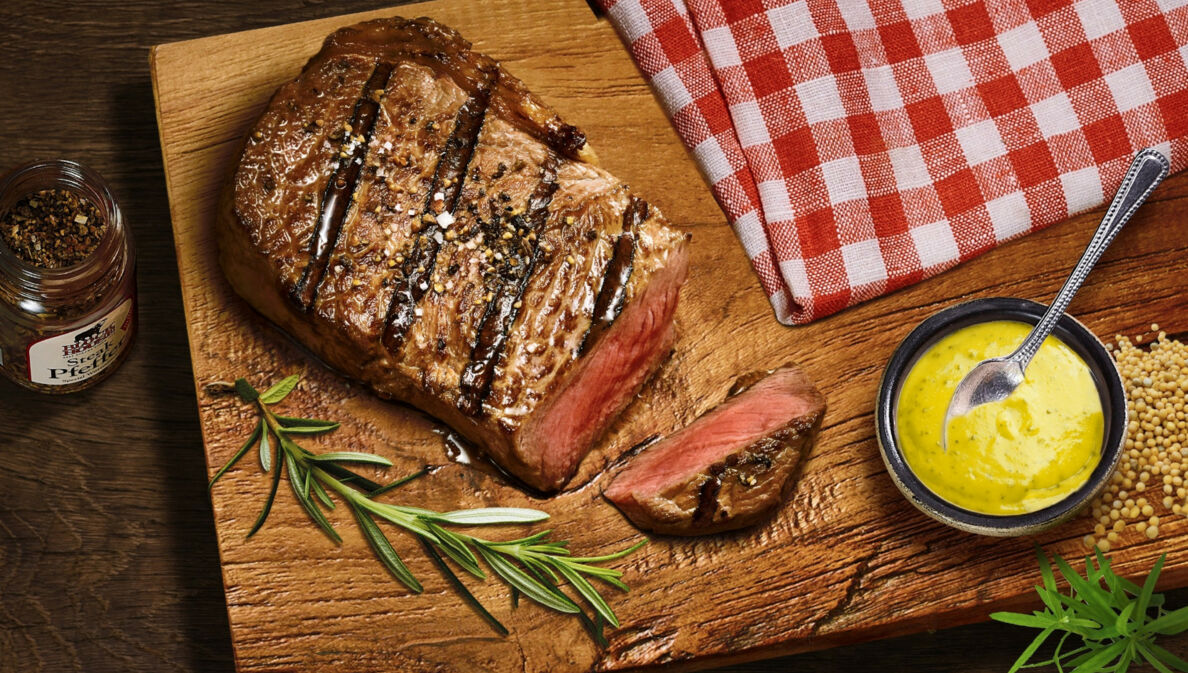 Aufsicht eines gebratenen, angeschnittenen Steaks auf einem Brett neben einer Schale mit Senf.