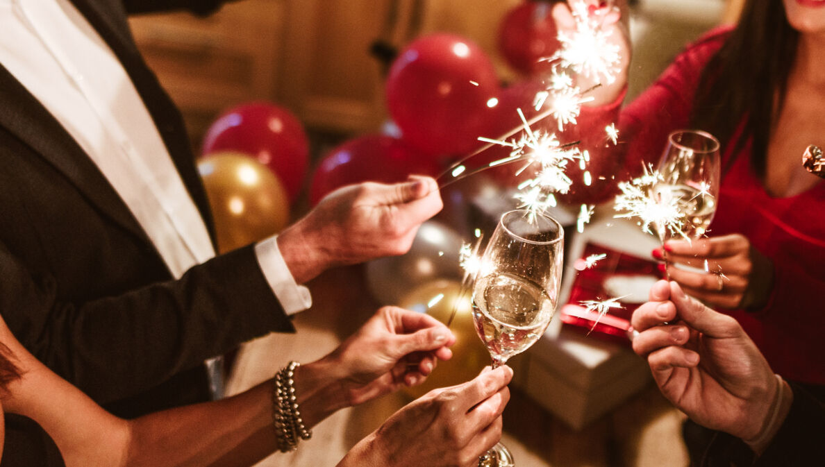 Nahaufnahme der Arme von vier edel gekleideten Personen mit Sektgläsern und Wunderkerzen im feierlichen Ambiente mit Luftballons im Hintergrund.